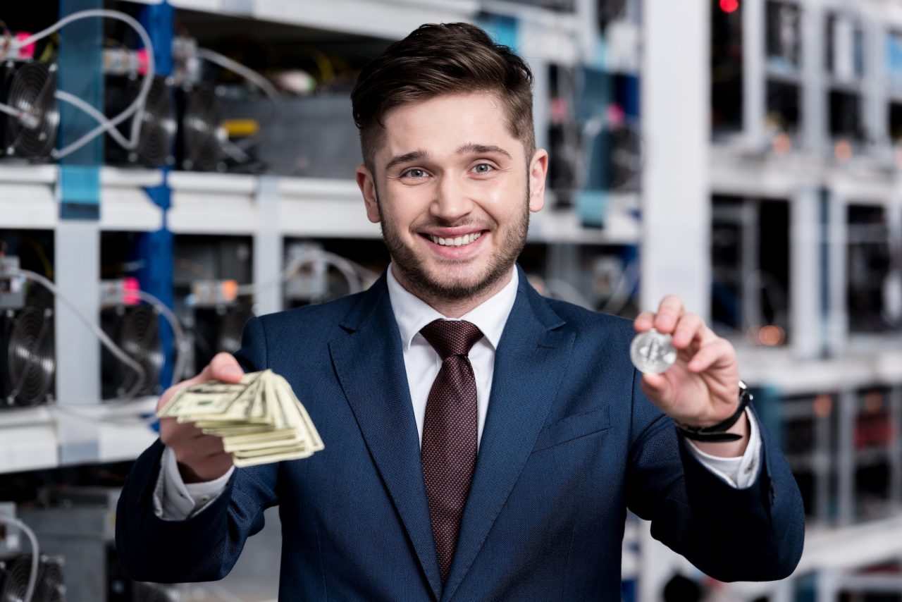 handsome young businessman holding stack of cash and bitcoin at cryptocurrency mining farm